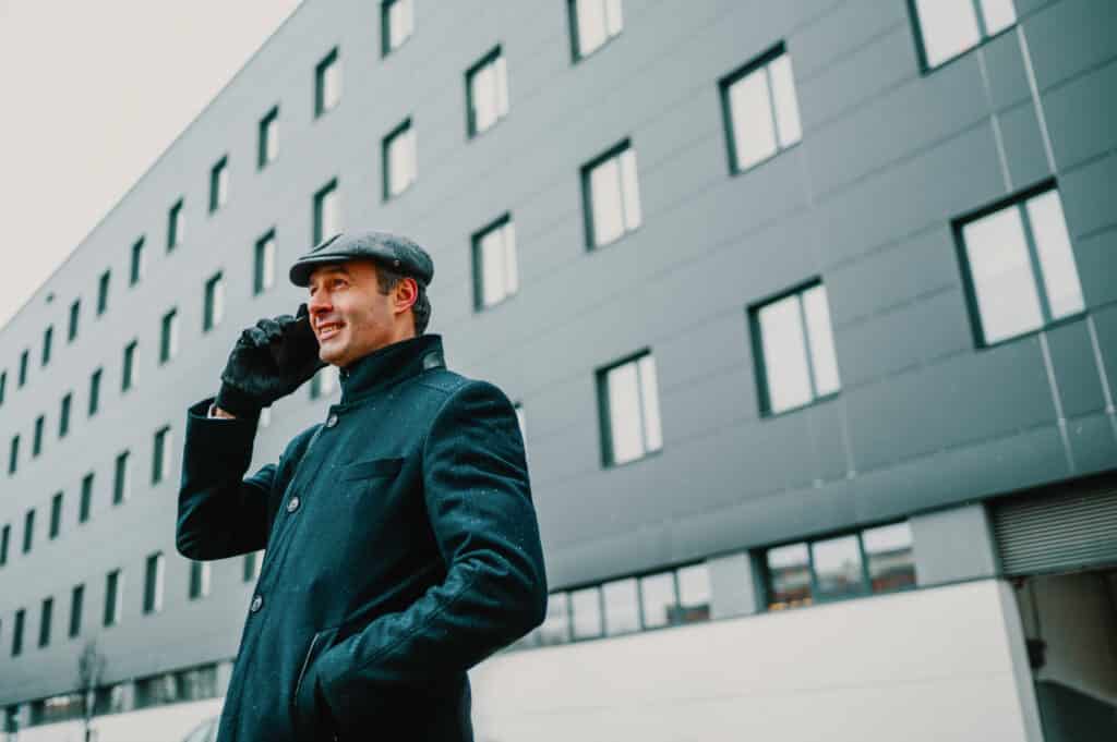 A cheerful trendy executive is standing in front of a corporate firm in a financial district and having a business phone call in rainy cold weather. An elegant businessman is talking on the phone.